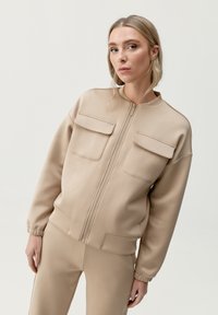 Beige zip-up bomberjacka i slätt material, med två bröstfickor och elastiska manschetter, ihop med matchande beige byxor.