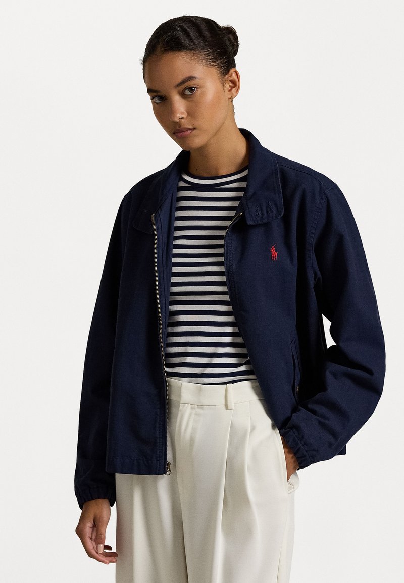 Marineblauwe jack met rits en kraag, voorzien van een klein rood logo, gedragen over een gestreept wit en marineblauw shirt, gecombineerd met beige high-waisted broeken.