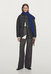 Giacca imbottita nera, sciarpa blu, camicia bianca, pantaloni grigi. Modello in piedi con guanti marroni, scarpe nere, sfondo neutro.