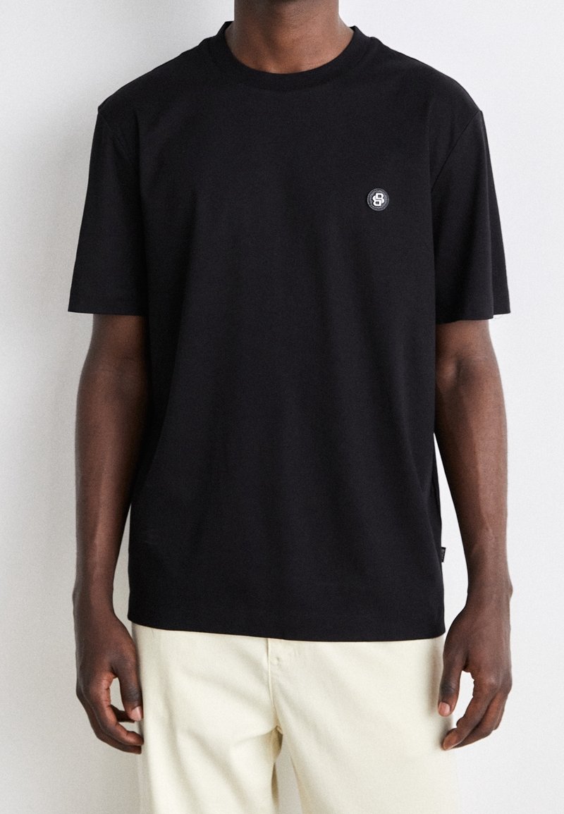 Homme portant un t-shirt noir à manches courtes avec un petit logo circulaire sur la poitrine gauche et un pantalon beige clair, debout devant un fond blanc.