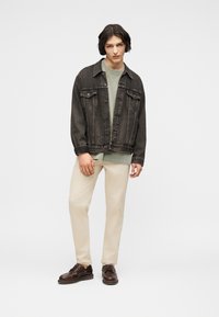 Jeune homme portant une veste en denim charbon, un sweat-shirt vert clair, un pantalon beige et des chaussures en cuir marron foncé, debout devant un fond blanc.