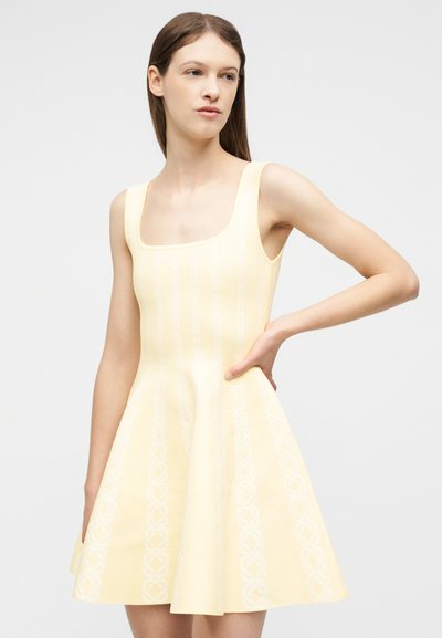 Νεαρή γυναίκα που φοράει αμάνικο, ανοιχτό κίτρινο φαρδύ φόρεμα με διακριτικά κάθετα σχέδια, στέκεται με το ένα χέρι στη μέση.