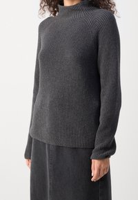 Femme portant un pull à col roulé côtelé gris foncé et une jupe noire, debout devant un fond uni clair.