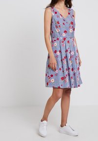 Vestido sin mangas con escote en V, con rayas azules y blancas y patrones florales en rojo y rosa. Diseño de falda acampanada, combinado con zapatillas blancas.