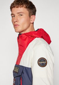Noor mees, kes kannab värvidega blokeeritud jakki, millel on punane kapuuts, valge ülemine osa ja tumesinine alumine osa. Jakil on varrukal ümmargune logo patch.