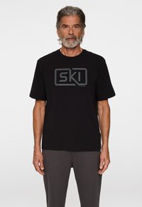 Mustat T-paita puuvillasta, jossa on harmaa "SKI" graafinen logo rinnassa. Lyhyet hihat, pyöreä pääntie ja rento leikkaus.