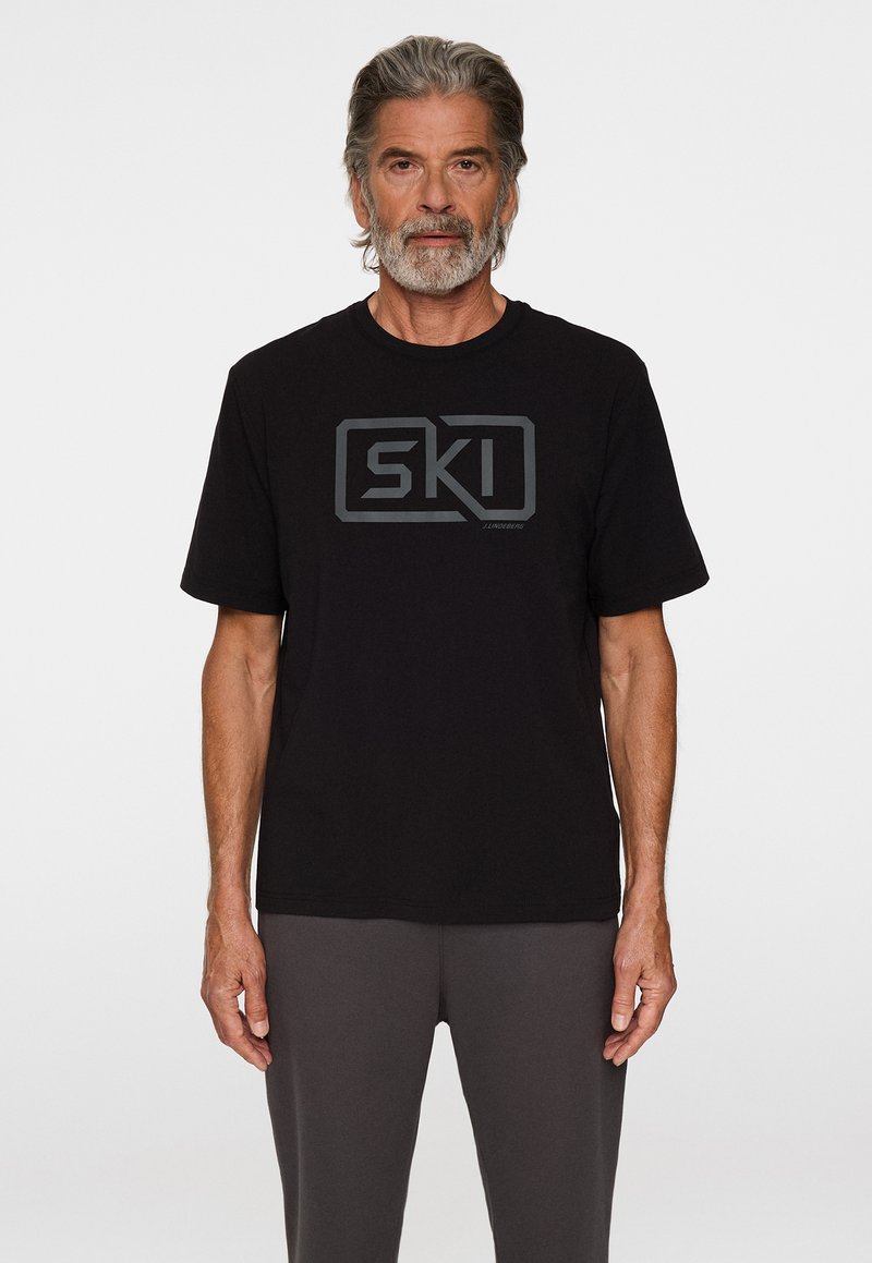 Mustat T-paita puuvillasta, jossa on harmaa "SKI" graafinen logo rinnassa. Lyhyet hihat, pyöreä pääntie ja rento leikkaus.