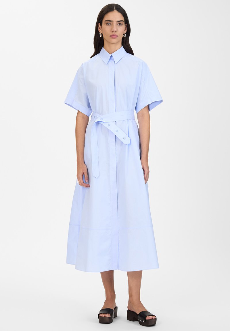 Abito camicia in cotone azzurro chiaro con maniche corte, un colletto, una cintura in vita aderente e una gonna ampia che scende sotto il ginocchio. Sandali neri con zeppa.