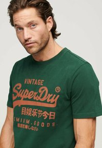 Uomo con capelli castani corti che indossa una maglietta verde con il logo "SuperDry Vintage" e testi in giapponese e inglese, su uno sfondo chiaro e semplice.