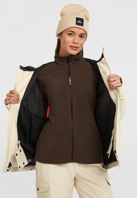 Bruine fleece binnenjack met hoge kraag, gedragen onder een crèmekleurig buitenjack. Beigekleurige gebreide beanie op persoon, minimalistisch ontwerp, geen patronen.