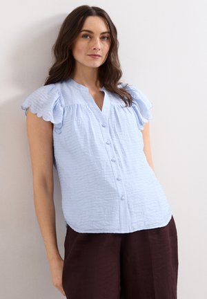 Vrouw met lang bruin haar draagt een lichtblauw gestructureerd blouseje met gegolfde korte mouwen en donkerbruine broek, staand tegen een witte muur.