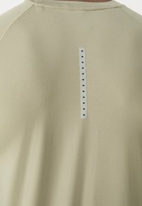 Camisa de un tono caqui claro con una textura suave, que presenta un tabulador vertical blanco con agujeros negros en la parte posterior, escote redondo y mangas raglán.