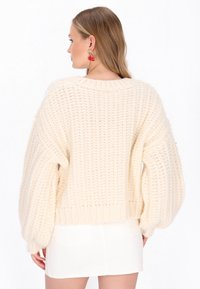 Krémový pletený sveter s voľným strihom, textúrovaným vzorom a širokými rukávmi. Skombinovaný s bielou mini sukňou, ktorá ukazuje uvoľnený štýl.