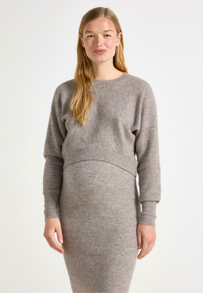 Donna sorridente con capelli lunghi che indossa un maglione grigio lavorato a maglia e un vestito abbinato, con le mani gentilmente appoggiate ai lati su uno sfondo chiaro.