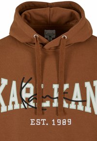 Sweat à capuche marron avec texte brodé en blanc et noir "KAPELKAN" et "EST. 1989,", avec cordons et étiquette intérieure visible.