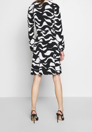 Černé šaty s dlouhým rukávem, ozdobené bílým abstraktním vlnkovým vzorem, s páskem v pase a rozšířenou sukní. Doplněno černými sandálemi na vysokém podpatku s pásky.