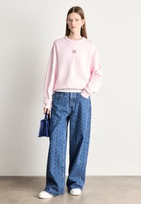 Różowy sweter z małym logo, w połączeniu z dżinsowymi spodniami o szerokich nogawkach z powtarzającym się wzorem tekstu. Białe sneakersy i niebieska torebka.