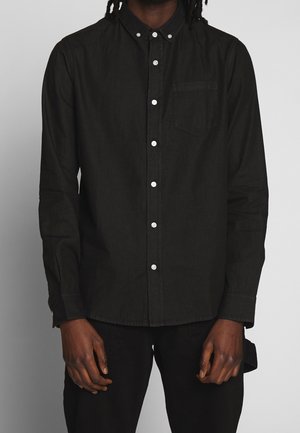 Mężczyzna ubrany w czarną koszulę z długim rękawem, białymi guzikami i kieszonką na piersi, stoi na jednolitym tle.