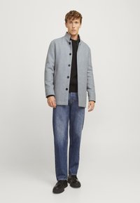 Abrigo de mezcla de lana gris claro con cuello alto, cierre de botones y tela texturizada, combinado con un suéter negro y jeans azules. Unos zapatos negros completan el look.
