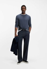 Camicia a maniche lunghe blu navy con colletto bianco, pantaloni ampi in denim scuro, sneakers nere e giacca blu navy appoggiata sul braccio.