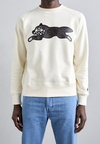 Uomo che indossa un sudore color crema con un grafico di un cane cartone animato marrone scuro con denti aguzzi, abbinato a jeans blu, su uno sfondo semplice.