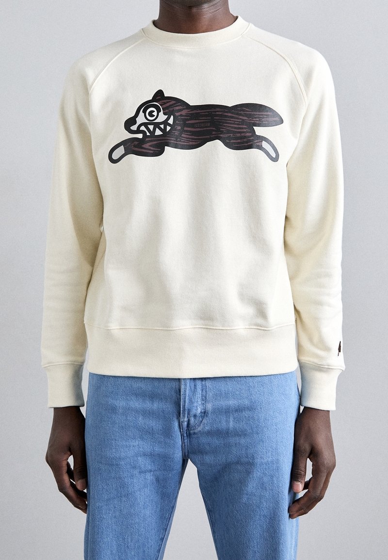 Uomo che indossa un sudore color crema con un grafico di un cane cartone animato marrone scuro con denti aguzzi, abbinato a jeans blu, su uno sfondo semplice.