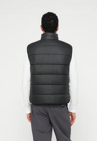 Melna pūkaina veste ar augstu apkakli, horizontāla izšūšanas struktūra, viegla materiāla, un aizmugures skats, kas rāda minimālu detaļu un logotipa birku.