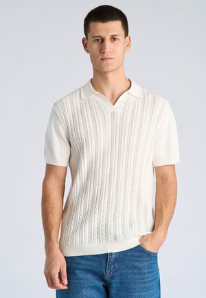 Jonge man met kort donker haar, gekleed in een wit gebreid poloshirt met korte mouwen en een blauwe spijkerbroek, staand met één hand in zijn zak.