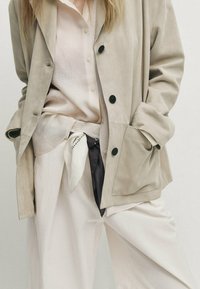 Beige mokkanakki, jossa on mustat napit, ylle puettuna vaaleanpunaisen paidan kanssa. Huomionarvoisia yksityiskohtia ovat valkoinen huivi mustilla korostuksilla vyötäröllä.