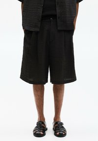 Pantaloni corti neri con una vestibilità ampia, dotati di tasche laterali. Abbinati a una camicia a maniche corte coordinata e sandali neri.