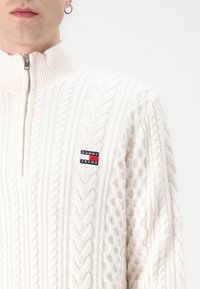 Wit kabelgebreid sweater met hoge kraag en ritssluiting. Bevat een kleine rood, wit en blauwe "Tommy Jeans" patch op de borst.
