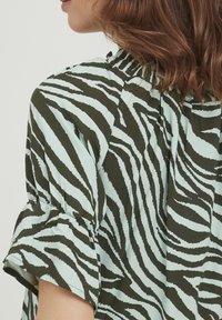 Camicia a maniche corte di colore azzurro chiaro con strisce zebra verde scuro, presenta un colletto arricciato e un orlo delle maniche leggermente arricciato.
