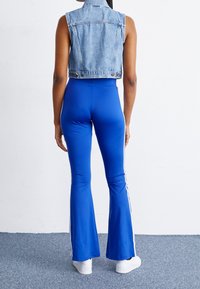 Chaleco de mezclilla recortado con un respaldo estructurado y un cuello tipo banda, combinado con pantalones ajustados acampanados azules que presentan rayas laterales blancas. Zapatillas blancas complementan el conjunto.