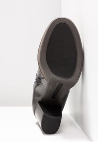 Chaussure en cuir noir avec un talon plat et une semelle en caoutchouc texturée. Présente des coutures discrètes et un design épuré. Étiquette de taille 37 visible.
