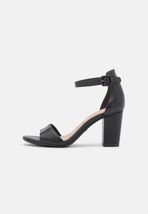 Sort hælsandal med en buet rem, åben tå og ankelspænde. Har en robust blokhæl og glat lædertekstur.