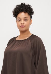 Brązowa, lekka bluzka z okrągłym dekoltem, marszczonymi detalami i luźnymi rękawami. Gładka tekstura z komfortowym krojem.