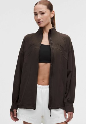 Vrouw met naar achter getrokken haar, die een donkerbruine ritsjas draagt over een zwarte sportbeha en witte korte broek tegen een effen achtergrond.