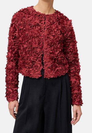 Veste rouge texturée à manches longues avec application florale 3D portée sur un pantalon noir taille haute à jambes larges sur une personne debout.