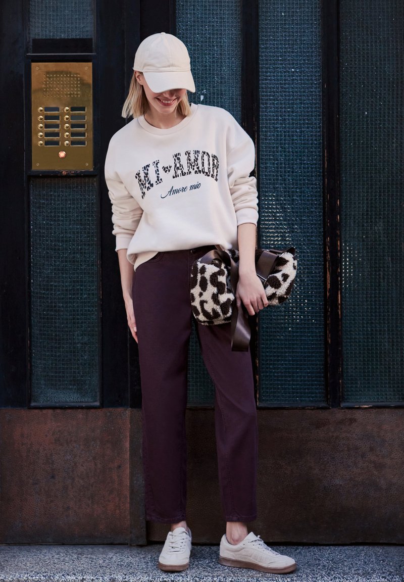 Femme portant une casquette beige clair, un sweat crème avec un texte, un pantalon foncé, des baskets blanches, tenant un sac à imprimé léopard, debout près d'une porte en bois sombre.