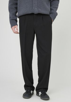 Pantalons noirs à jambe droite, avec des plis subtils et une texture lisse. Associés à un cardigan en maille gris et des baskets foncées.
