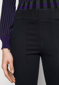 Černé džíny mají hladkou texturu s kontrastním modrým prošíváním. Vysoký pas zaručuje těsný střih, který doplňuje žebrovaný fialový svetr.