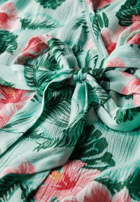 Bloemenwrap met een mintgroene achtergrond, grote roze bloemen en groene bladeren, met een gestrikt knoopdetail in de stof. Zachte, lichtgewicht textuur.