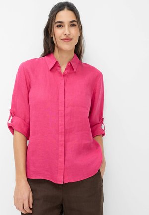 Młoda kobieta o długich ciemnych włosach, ubrana w jaskraworóżową koszulę na guziki z podwiniętymi rękawami i ciemnobrązowe spodnie, na jasnym tle.