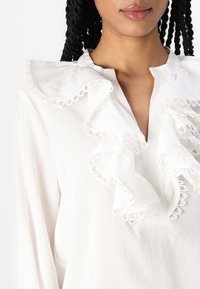 Hvit bluse med flosset krage og kant, med løkkedetaljer, lange ermer og knappelukking i halsen. Myk stofftekstur.