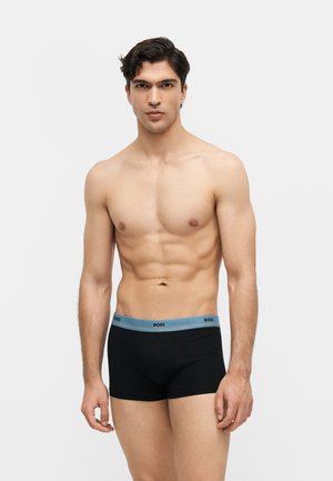 Muscular man standing against plain background wearing black boxer briefs with blue waistband labeled "BOSS."
