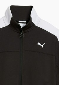 Blouson noir zippé avec col montant, accents blancs aux épaules et logo Puma blanc sur la poitrine gauche. Tissu texturé, design sportif.