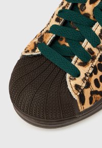 Superga z rjavim gumijastim koničkom prsta, leopardjim potiskom iz krzna na straneh in temno zelenimi vezalkami na belem ozadju.