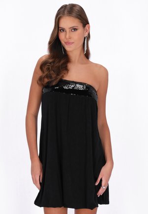 Vrouw met lang golvend bruin haar, gekleed in een mouwloze zwarte jurk met een pailletten halslijn en oorbellen in de vorm van een hart die bungelen.