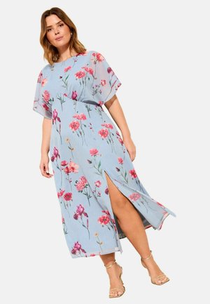 Vrouw draagt een lichtblauwe bloemenjurk met korte mouwen en een voorsplit, gecombineerd met gouden hoge hak sandalen met bandjes, staand op een witte achtergrond.