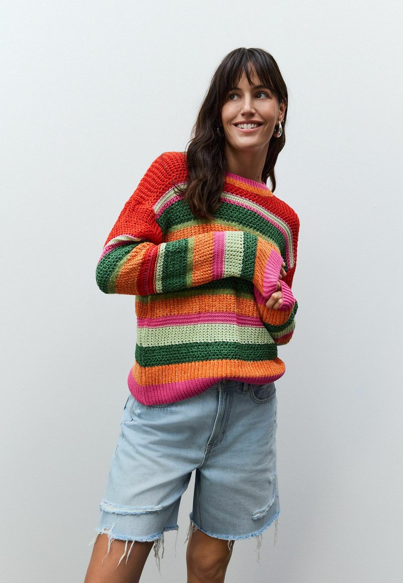Färgglad stickad tröja med horisontella ränder i röd, grön, rosa och orange; lös passform, rundad halsringning och långa ärmar. Bärs med ljusa denimshorts.